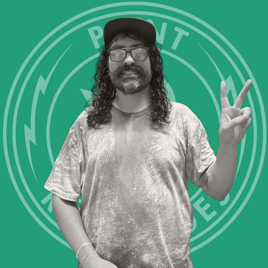 Man with long curly hair and beard wearing glasses, a cap, and a tie-dye shirt making a peace sign with his right hand.