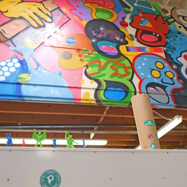 Colorful mural with abstract faces and shapes painted on a wall above a row of small green and blue plastic toy figures on a white surface.