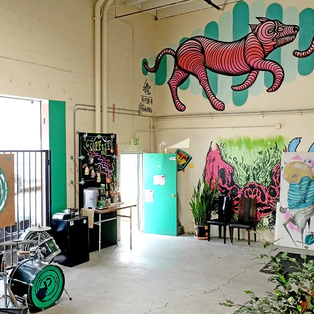 Interior of a creative cafe with a drum set, coffee bar, teal door, potted plants, and a colorful mural of a pink striped animal on the wall.