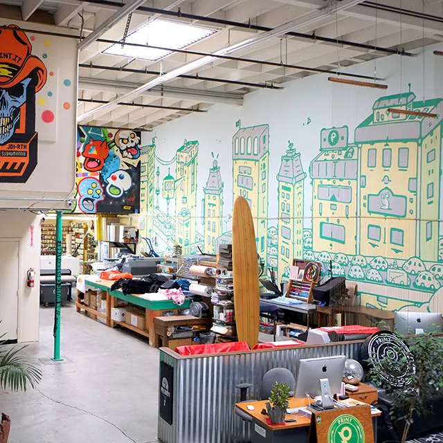 Modern creative workspace with mural of cartoon-style cityscape on wall, surfboard, and desks with computers and art supplies.