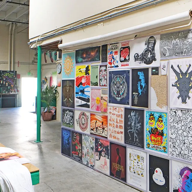 Indoor art gallery wall displaying various colorful framed artworks and posters in a bright, spacious room with a green pillar and exposed ceiling pipes.