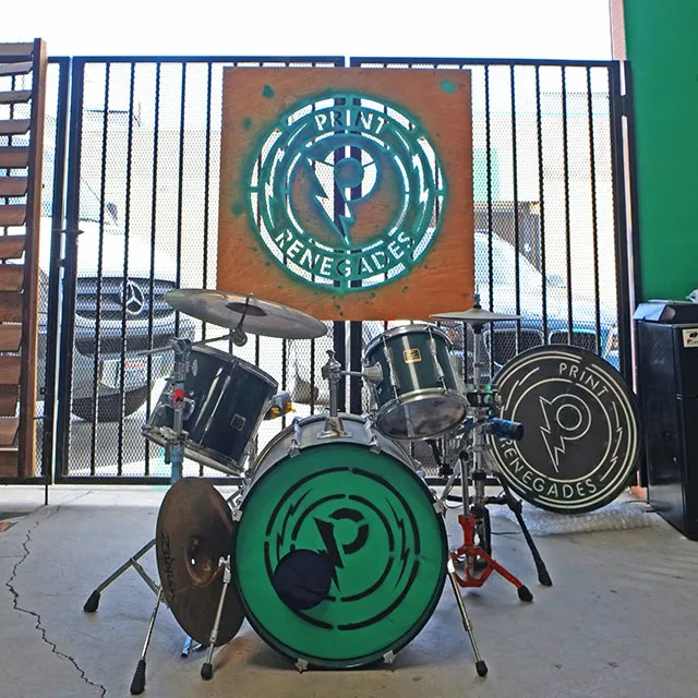 Drum set with Print Renegades logos on bass drum heads and a wooden sign with the same logo hanging on a metal gate in the background.