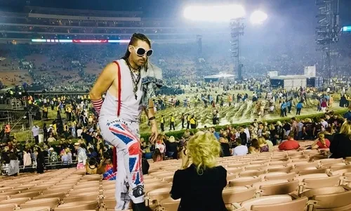 Man in white tank top, Union Jack pants, and large sunglasses posing in a stadium with a crowd and stage lights in the background.