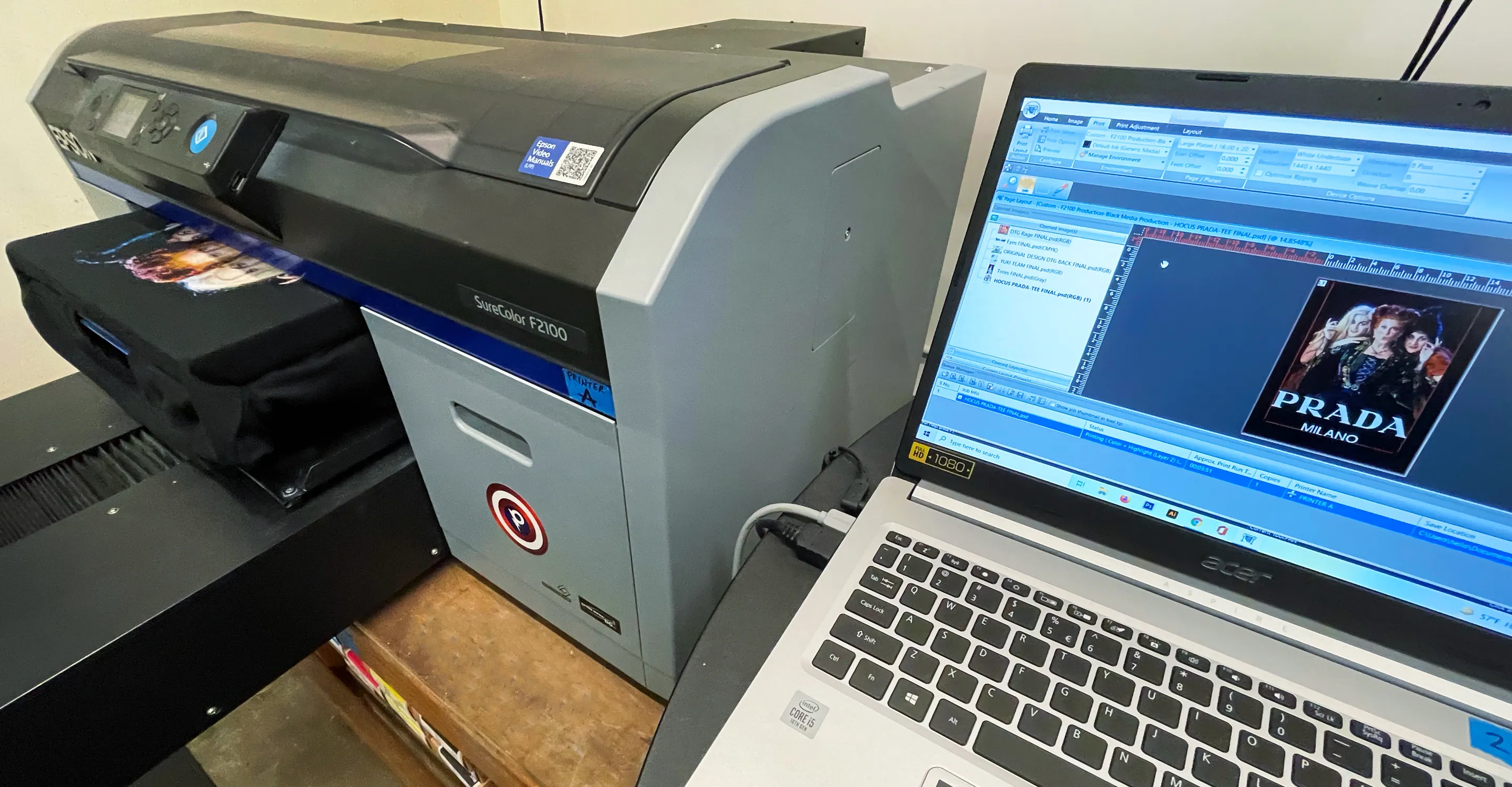 A SureColor F2100 direct-to-garment printer printing an image on a black t-shirt next to a laptop displaying the printing software with a Prada Milano design.