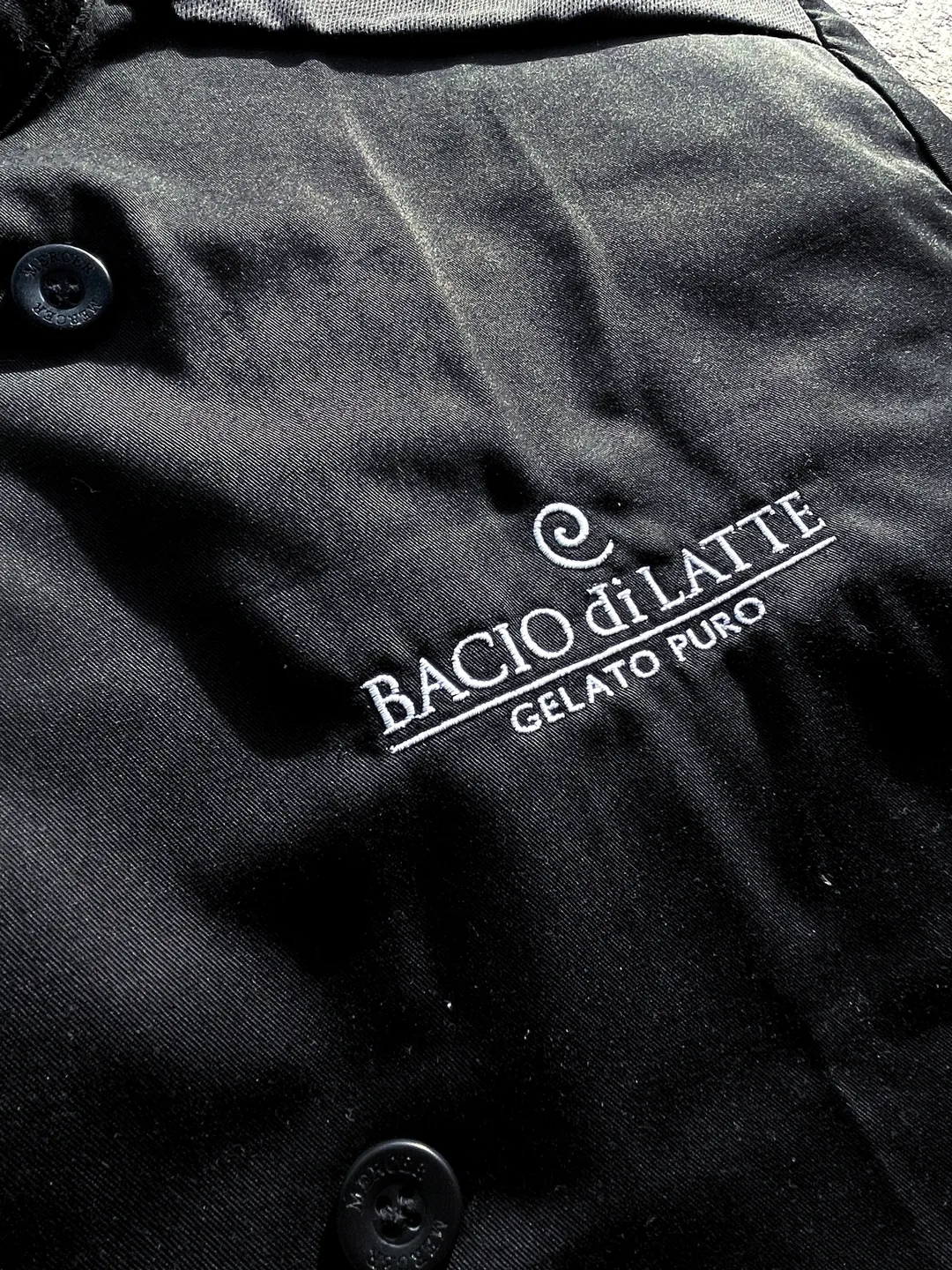 Close-up of black fabric with white embroidered text 'Bacio di Latte Gelato Puro' and two black buttons.