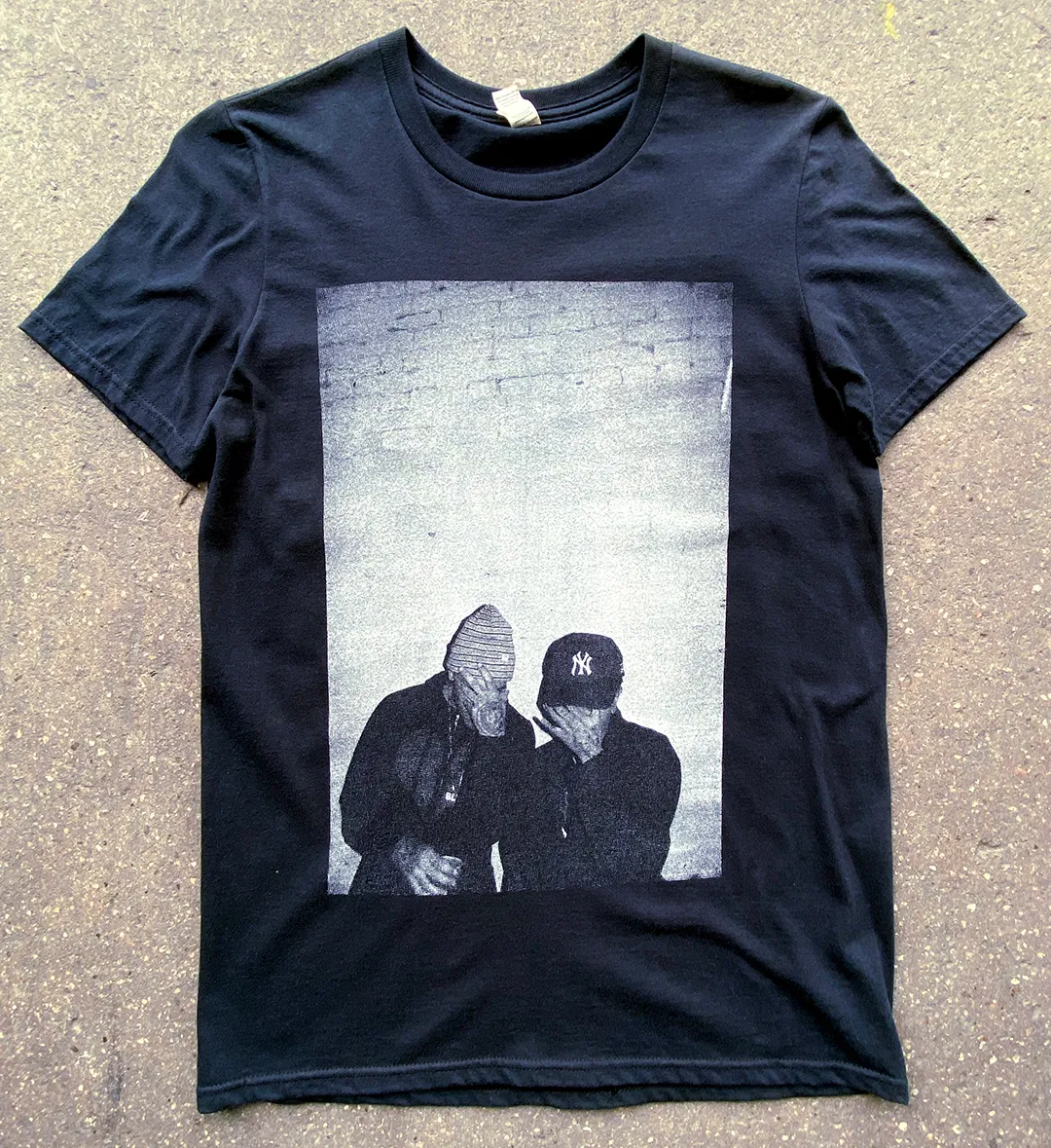 Black t-shirt with a grayscale print of two men covering their faces, one wearing a New York Yankees cap and the other a beanie.