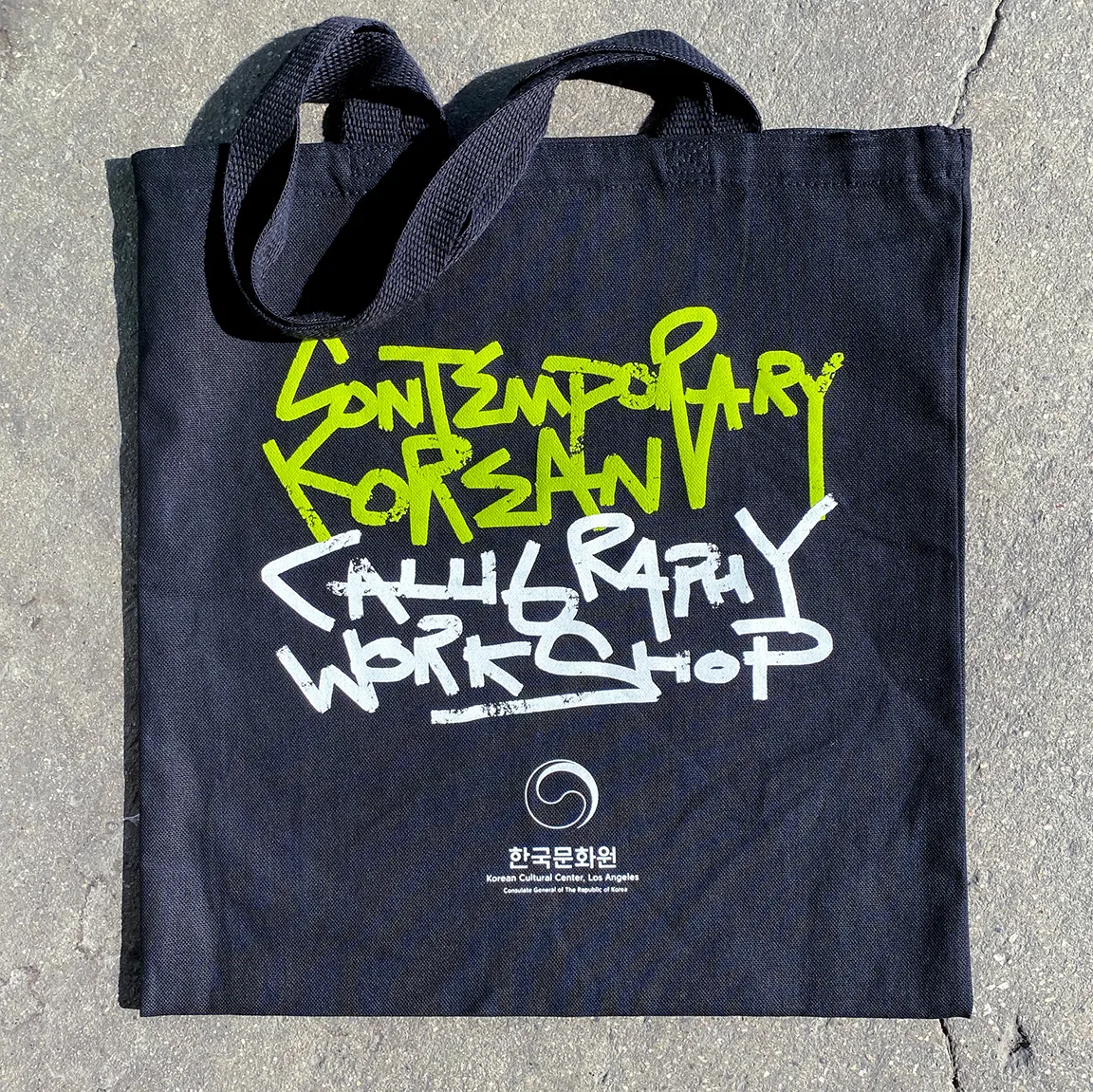 Black tote bag with green and white graffiti-style text reading 'Contemporary Korean Calligraphy Workshop' and a logo for the Korean Cultural Center, Los Angeles.
