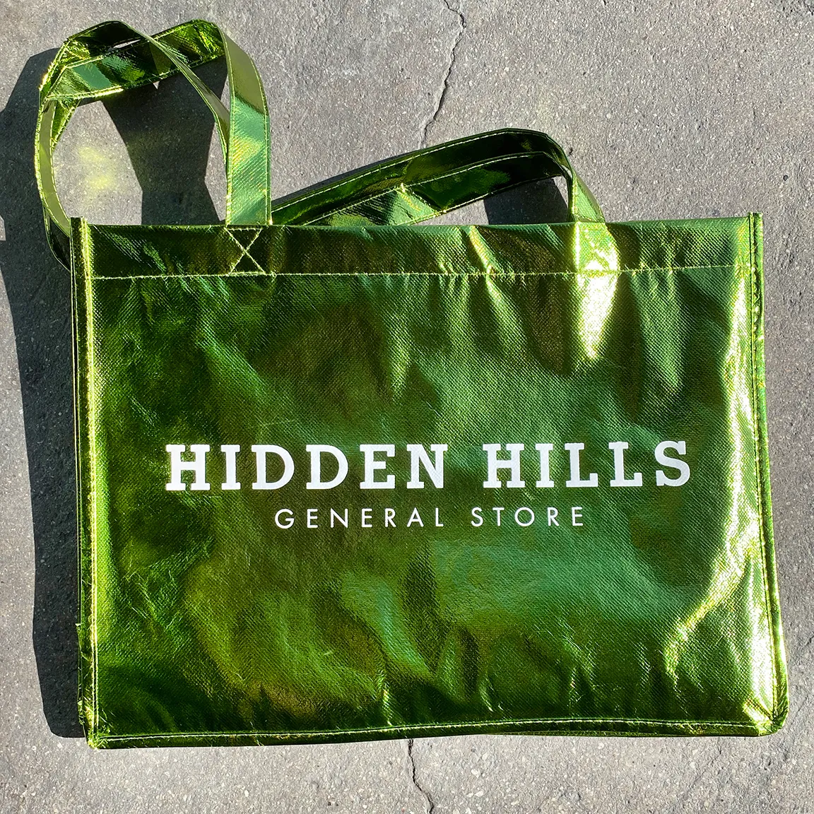 Shiny metallic green tote bag with white text reading Hidden Hills General Store on a concrete surface.