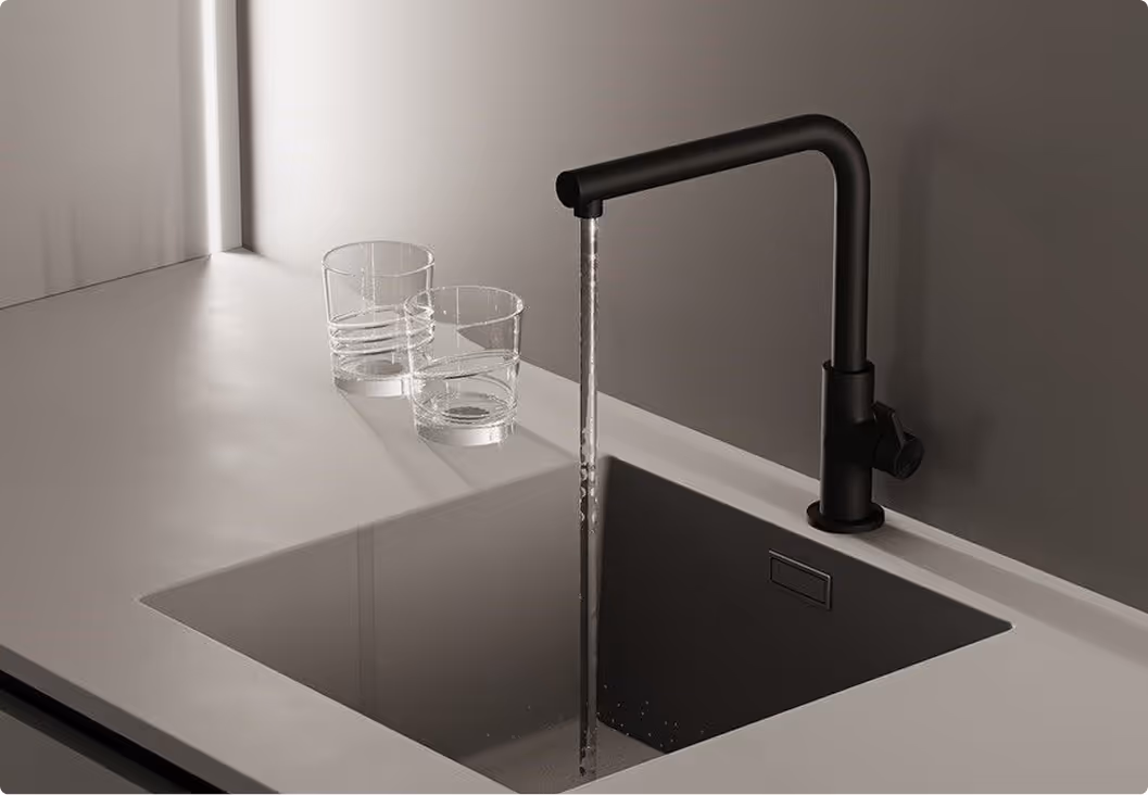Grifo negro sobre fregadero gris con agua corriente y dos vasos transparentes en la encimera.