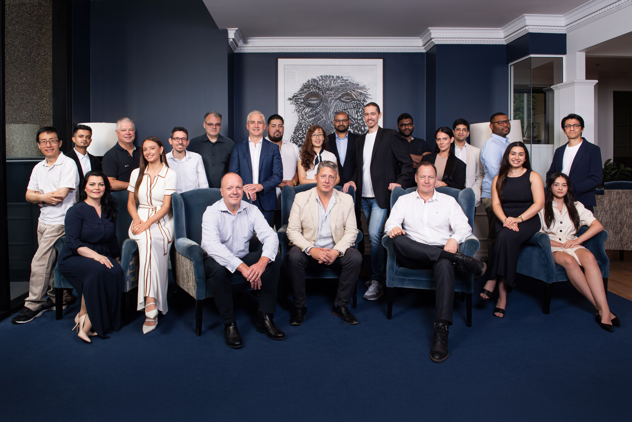 Group photo of 21 diverse professionals dressed in business casual attire posed in a modern office with navy walls and blue chairs.