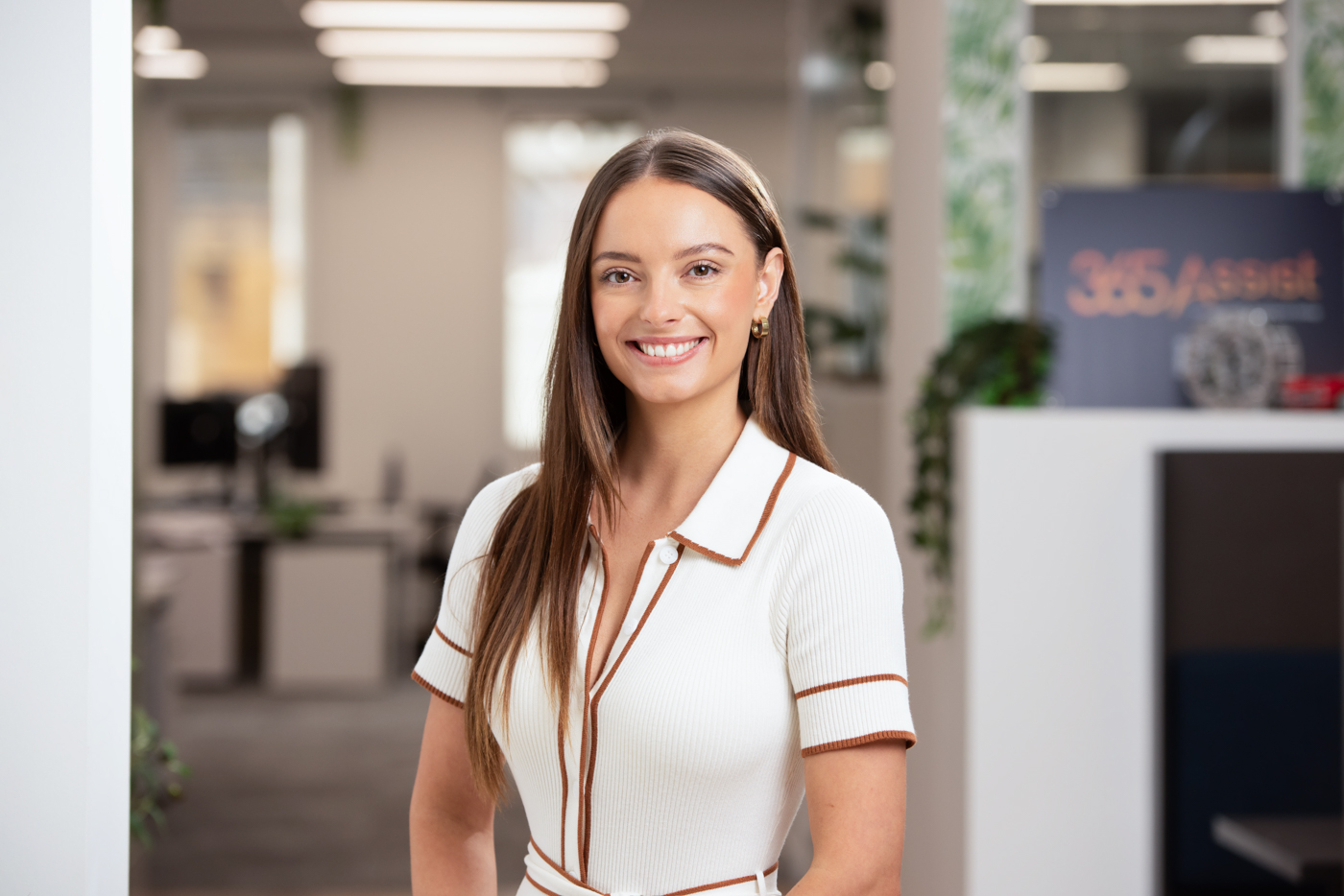 Smiling woman with long brown hair wearing a white dress with brown trim standing in a modern office.
