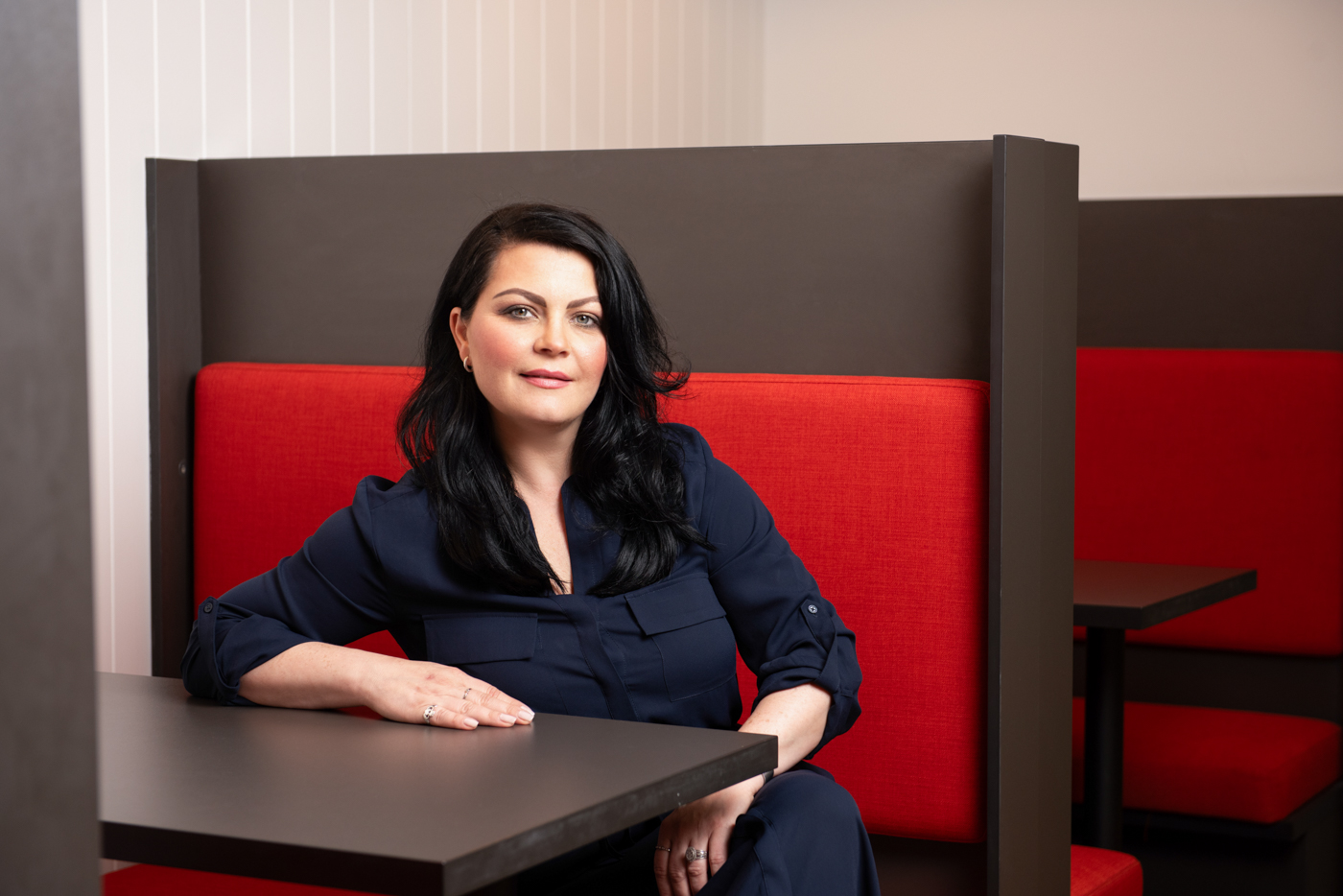 Woman with black hair wearing a navy blouse sitting at a table in a booth with red cushions.