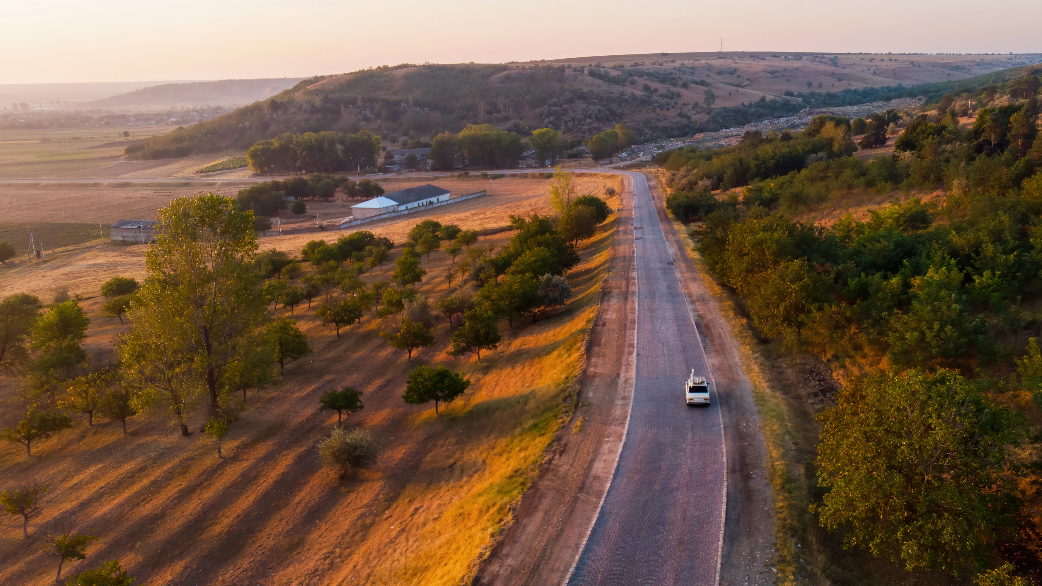 Car driving on a rural road surrounded by trees, fields, and hills at sunset.
