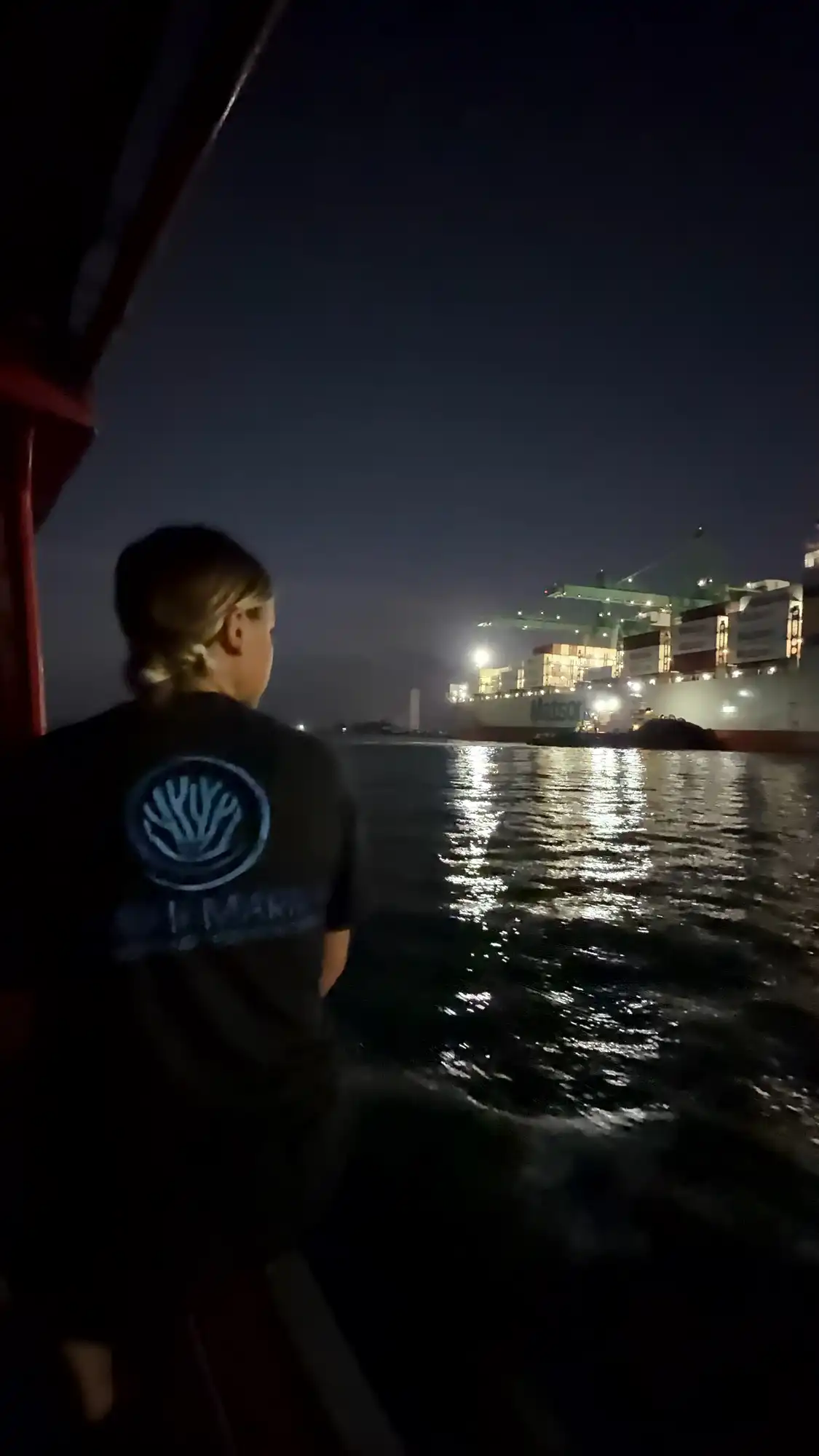 A Hawaiʻi MERC team member watches from a research boat as they pass a brightly lit container ship and port cranes at night in Guam.