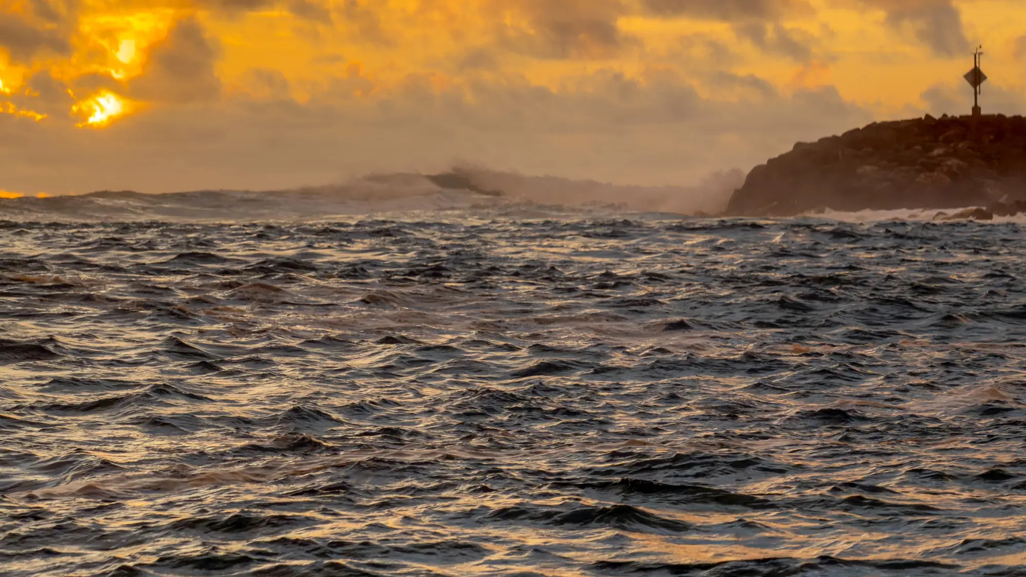Rough, choppy waters at sunset with waves crashing against a rocky breakwater in Guam during Typhoon Bavi weather conditions.