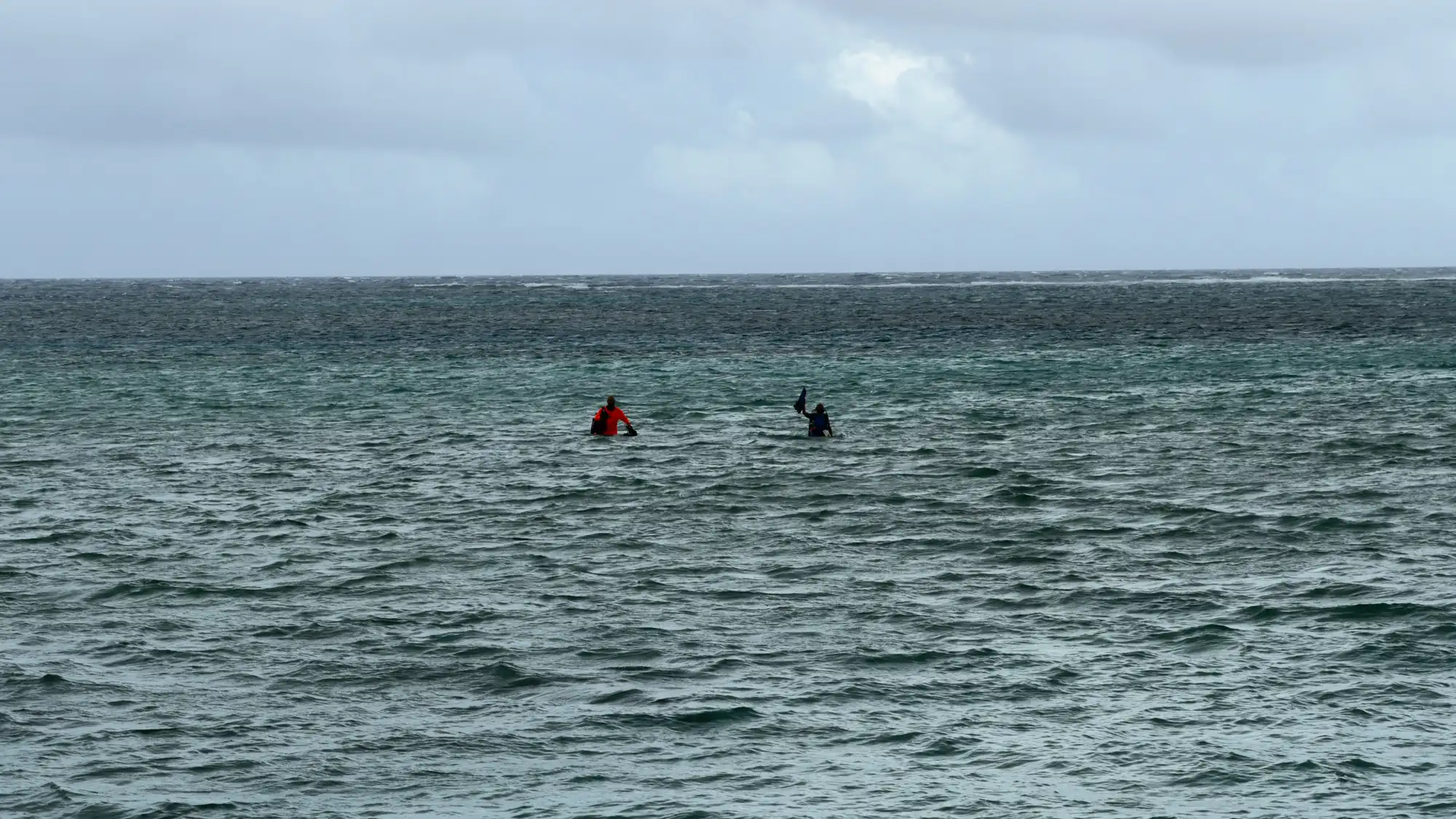 Two Hawaiʻi MERC divers in the ocean, one wearing bright orange, manage equipment during choppy, overcast conditions on the GAO Pacific Mission in Guam.