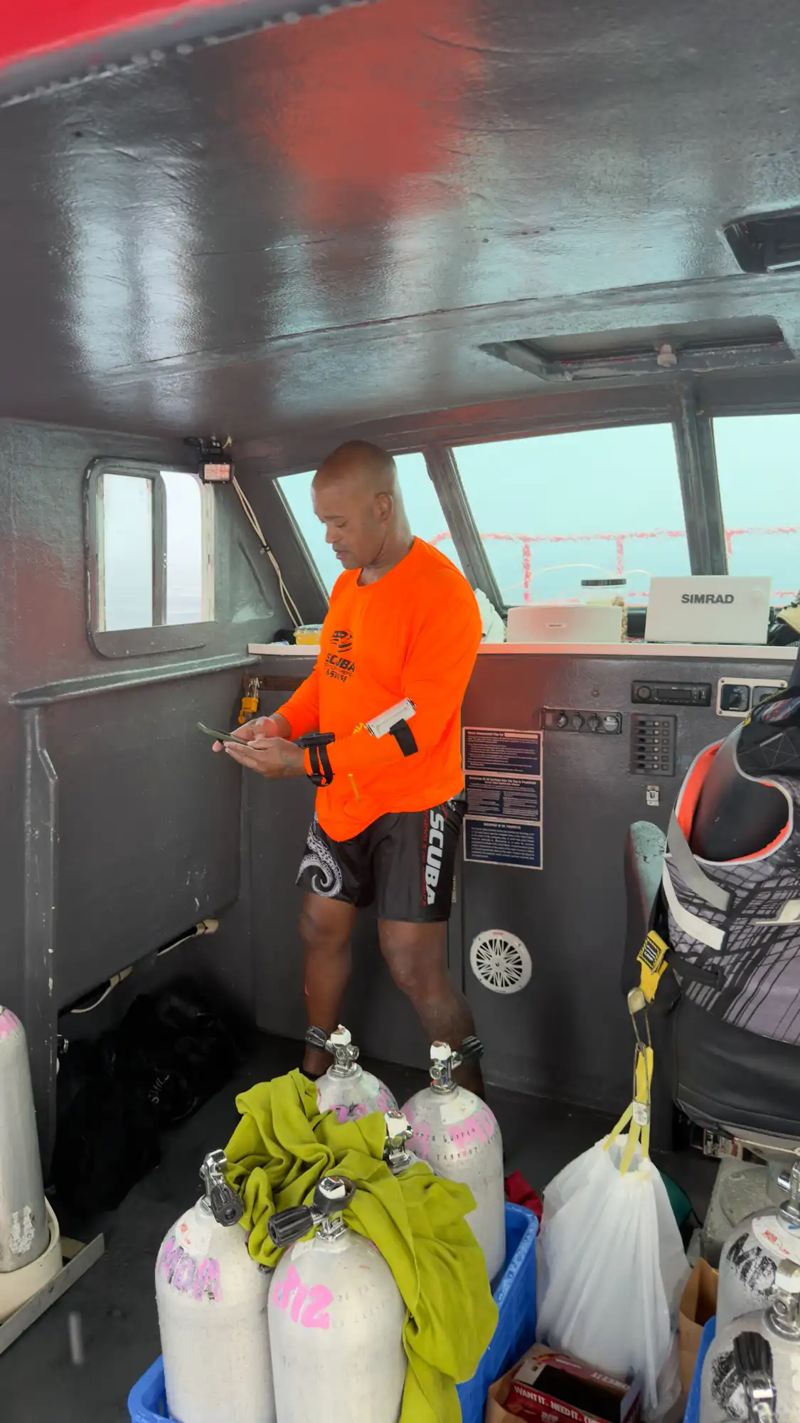 A Hawaiʻi MERC crew member in a bright orange shirt checks his phone inside a well-equipped research vessel cabin with scuba tanks in Guam.