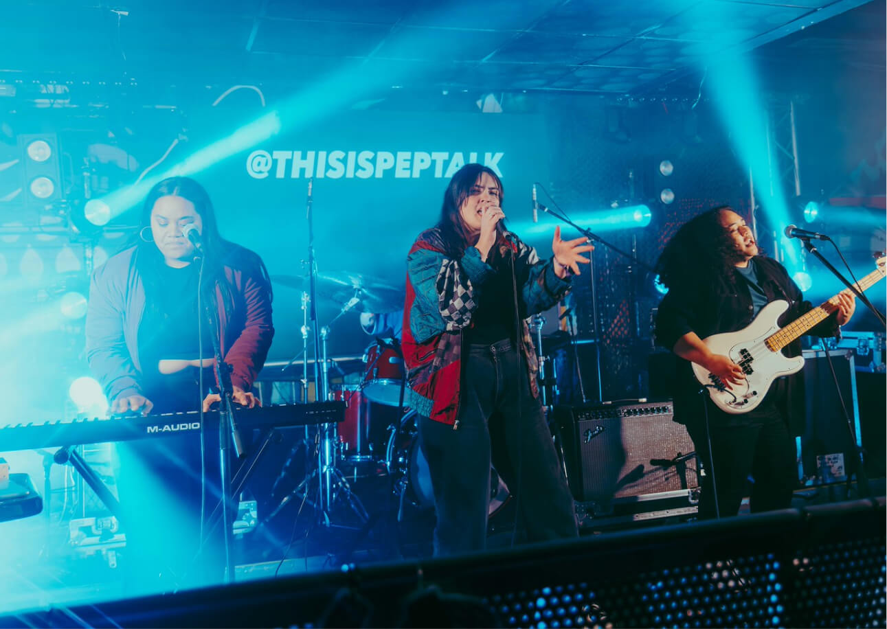 Three musicians performing on stage with keyboard, vocal microphone, and electric bass guitar under blue stage lights.