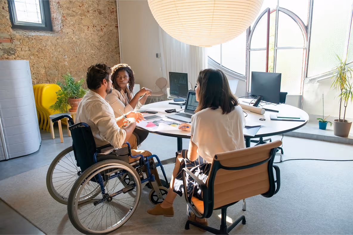 3 people in an office setting are talking - working together. One of them is on a wheelchair.