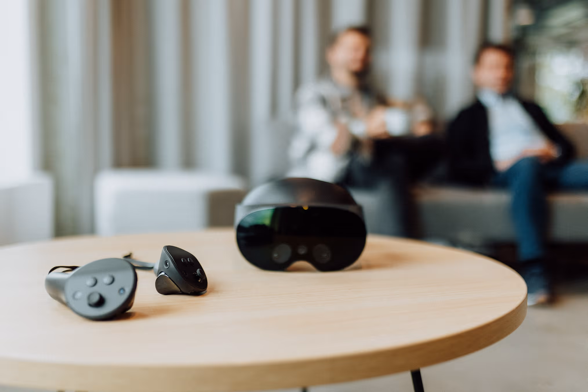 In the foreground there is a headset and controllers on a table. In the background there are people in office attire.