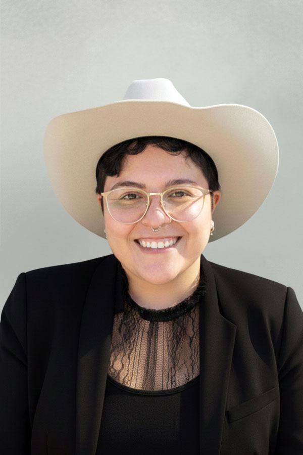 Smiling person wearing a white wide-brimmed hat, glasses, a septum ring, and a black blazer over a lace top.