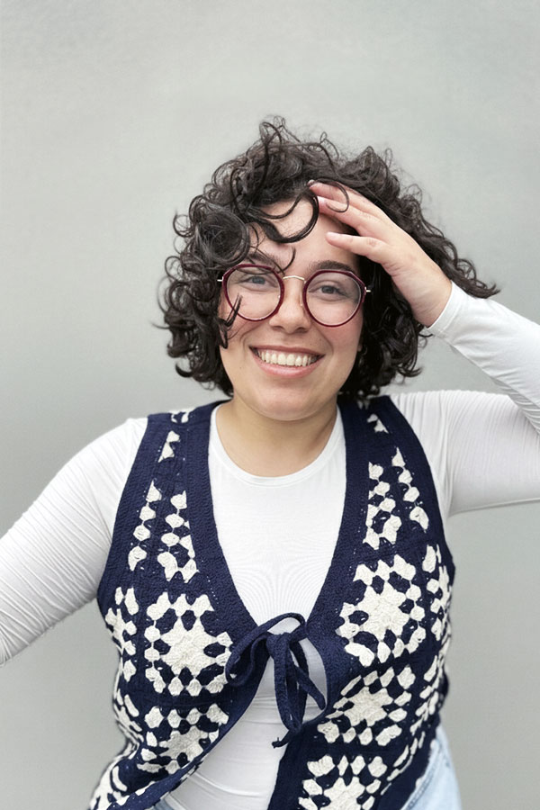 Smiling woman with curly hair and round glasses wearing a navy blue and white patterned vest over a white long-sleeve shirt, touching her head.
