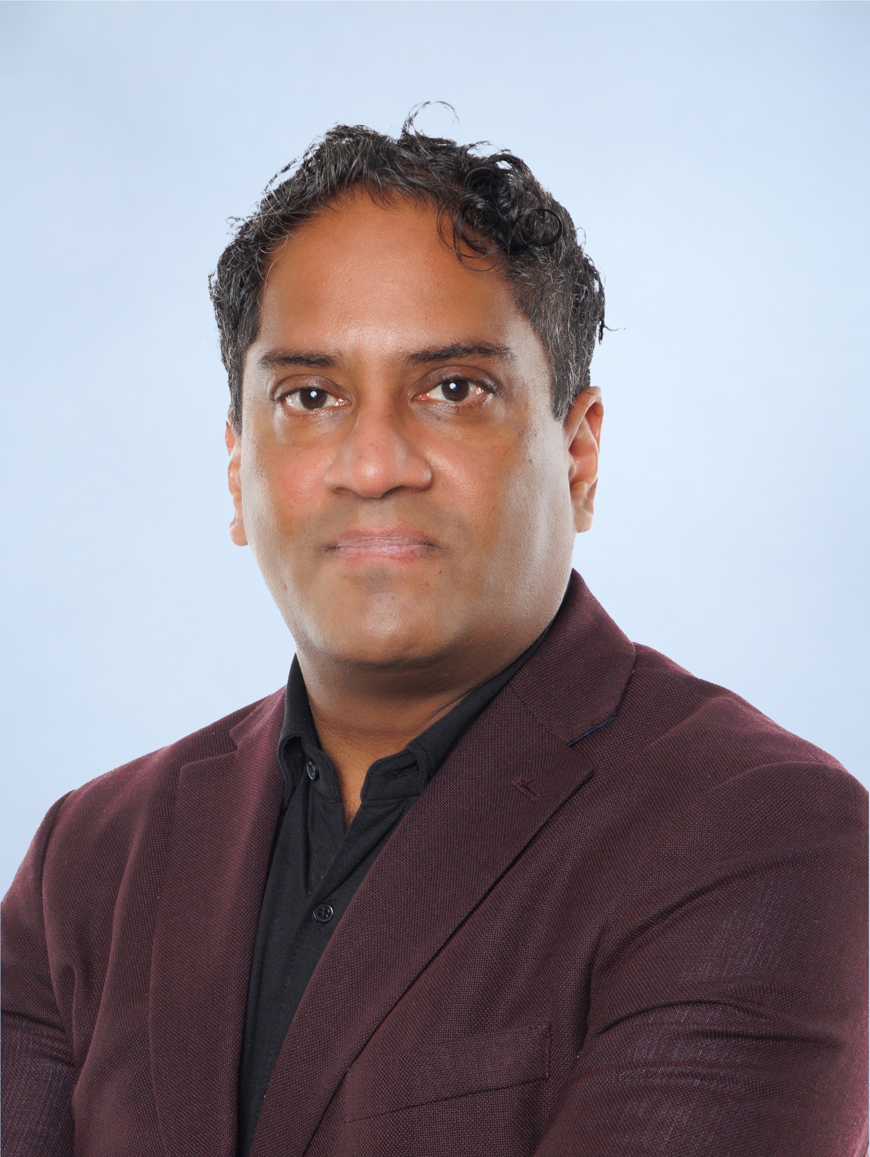 Portrait of a man with curly dark hair wearing a maroon blazer and black shirt against a light blue background.