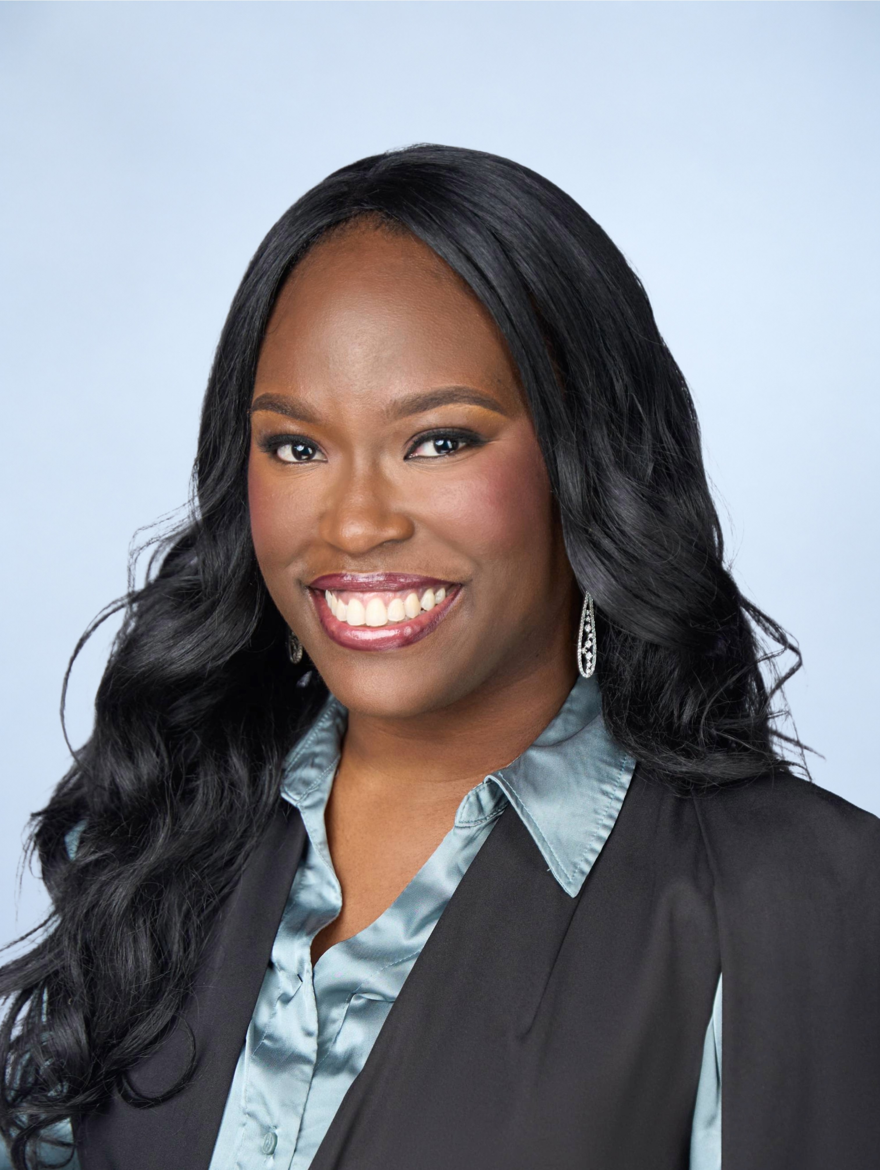 Smiling woman with long black wavy hair wearing silver earrings, a satin light blue blouse, and a black blazer, against a light blue background.