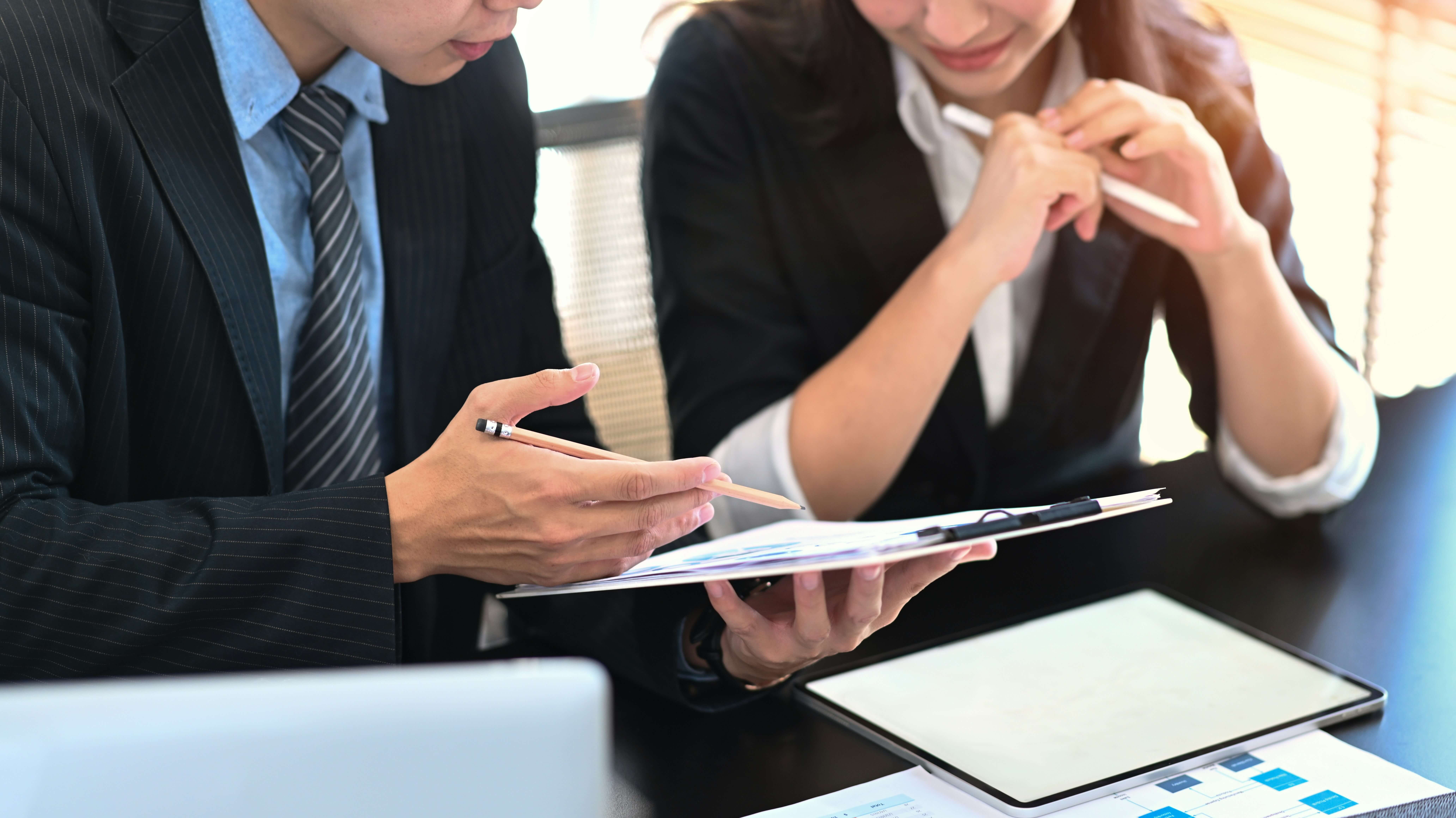 Two business professionals reviewing documents and charts during a meeting at a desk with a tablet and laptop.