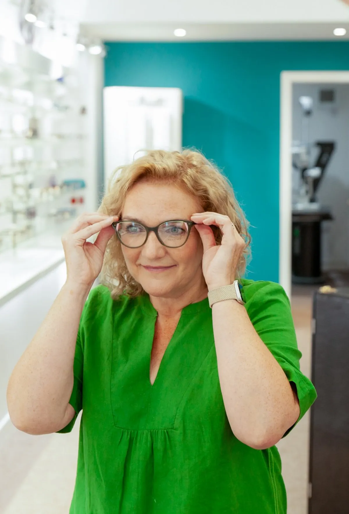 Middle-aged woman with curly blonde hair wearing a green top trying on eyeglasses in an optical store.