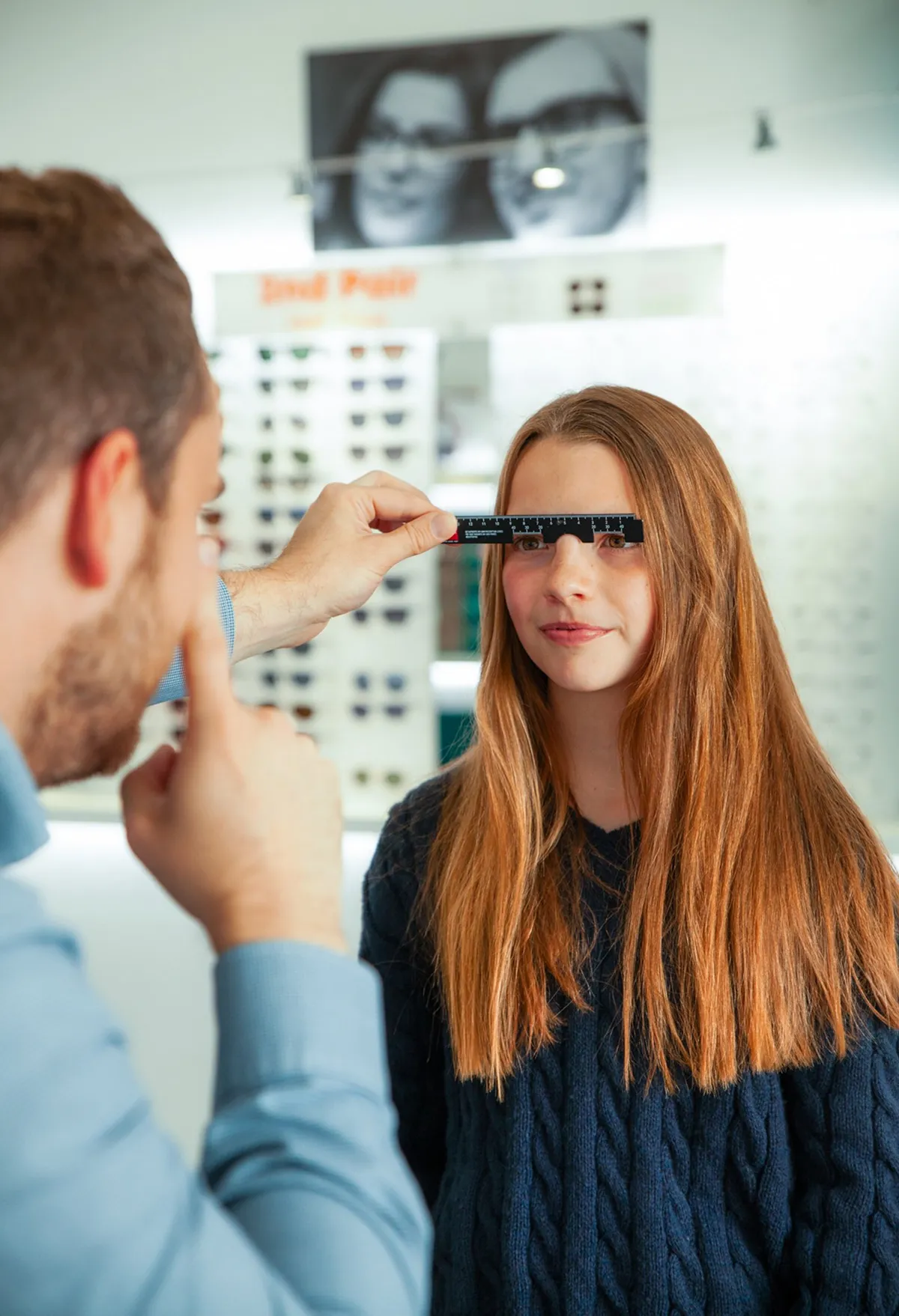 Optometrist measuring a young woman's pupillary distance with a ruler in an optical store.