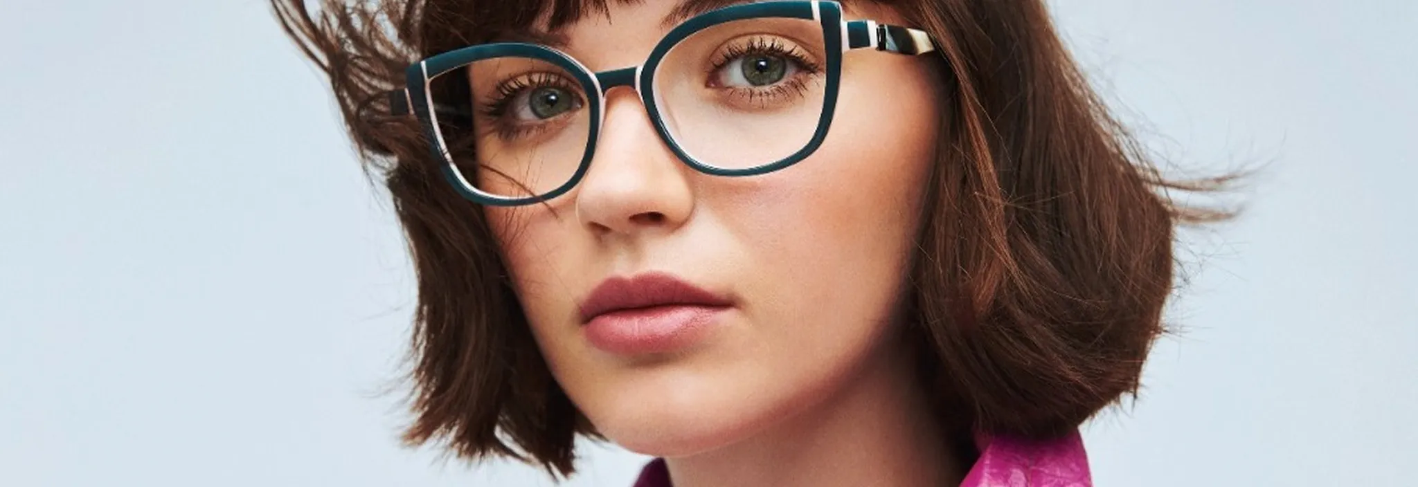 Close-up of a woman with short brown hair wearing large blue-framed glasses and a pink shirt.