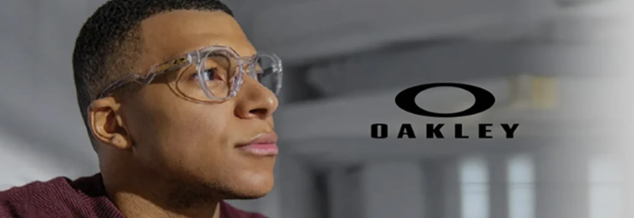 Man wearing clear round Oakley eyeglasses looking to the side with Oakley logo in the background.