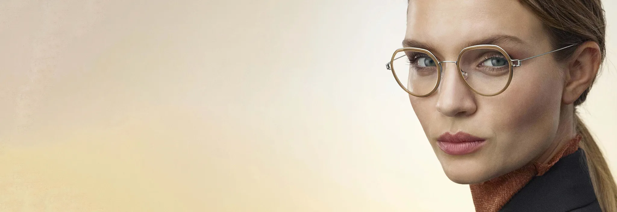 Close-up of a woman wearing round gold-rimmed glasses with a neutral expression and a beige background.