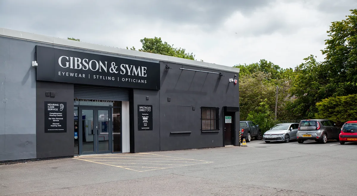 Exterior of Gibson & Syme opticians with a black facade and multiple parked cars nearby under a cloudy sky.