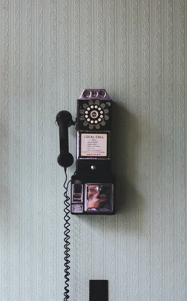 Vintage black rotary payphone mounted on a light green textured wallpaper wall.