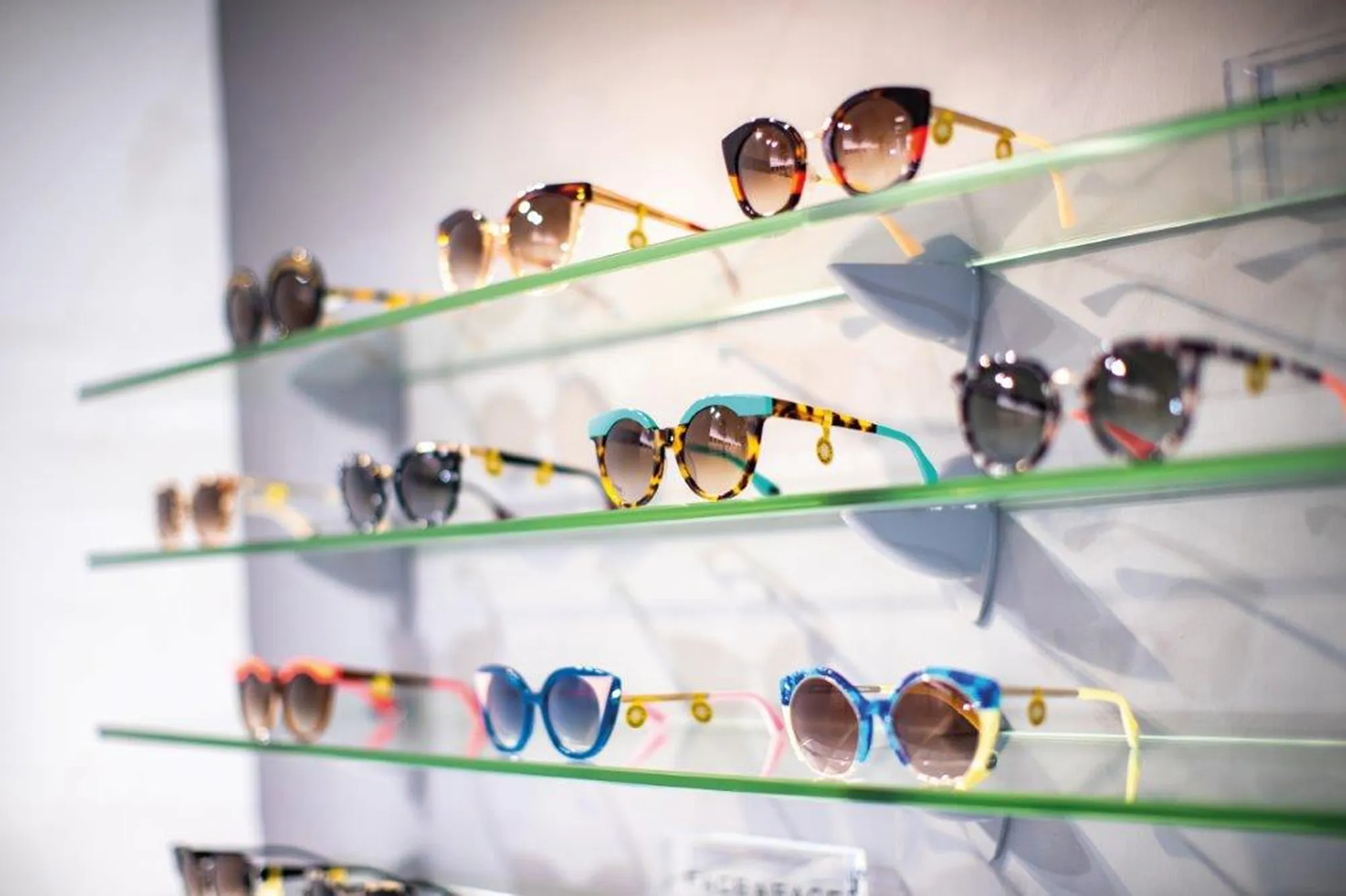 Various stylish sunglasses displayed on three glass shelves against a white wall in a retail store.