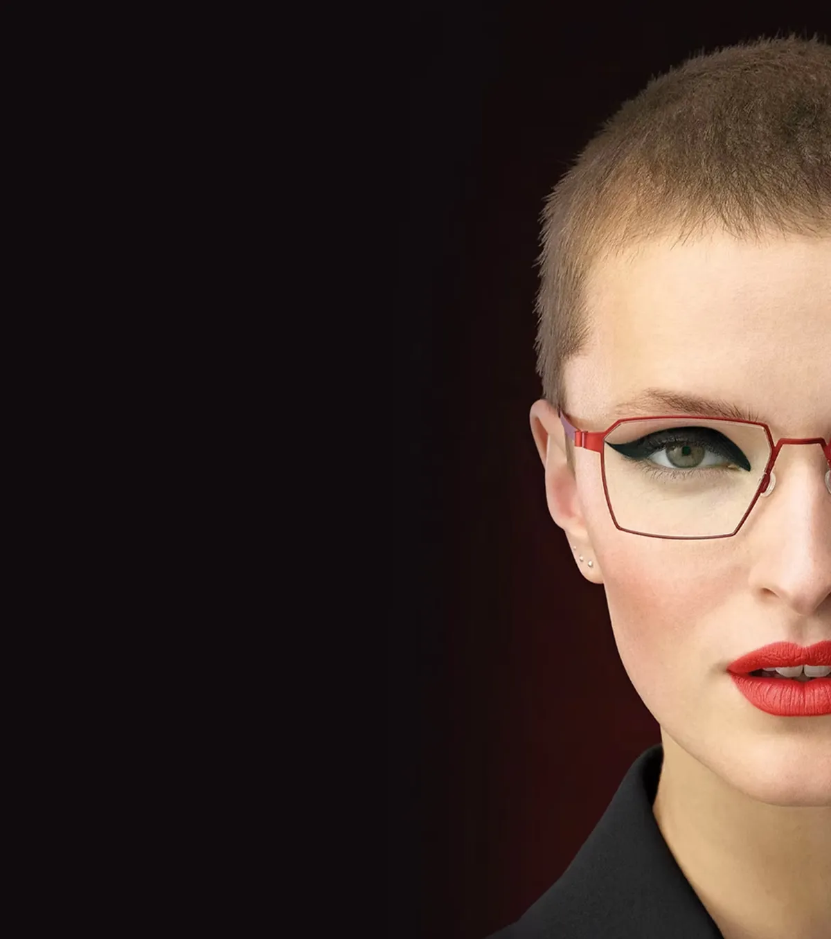 Portrait of a person with short hair wearing red glasses, bold eyeliner, and red lipstick against a dark background.