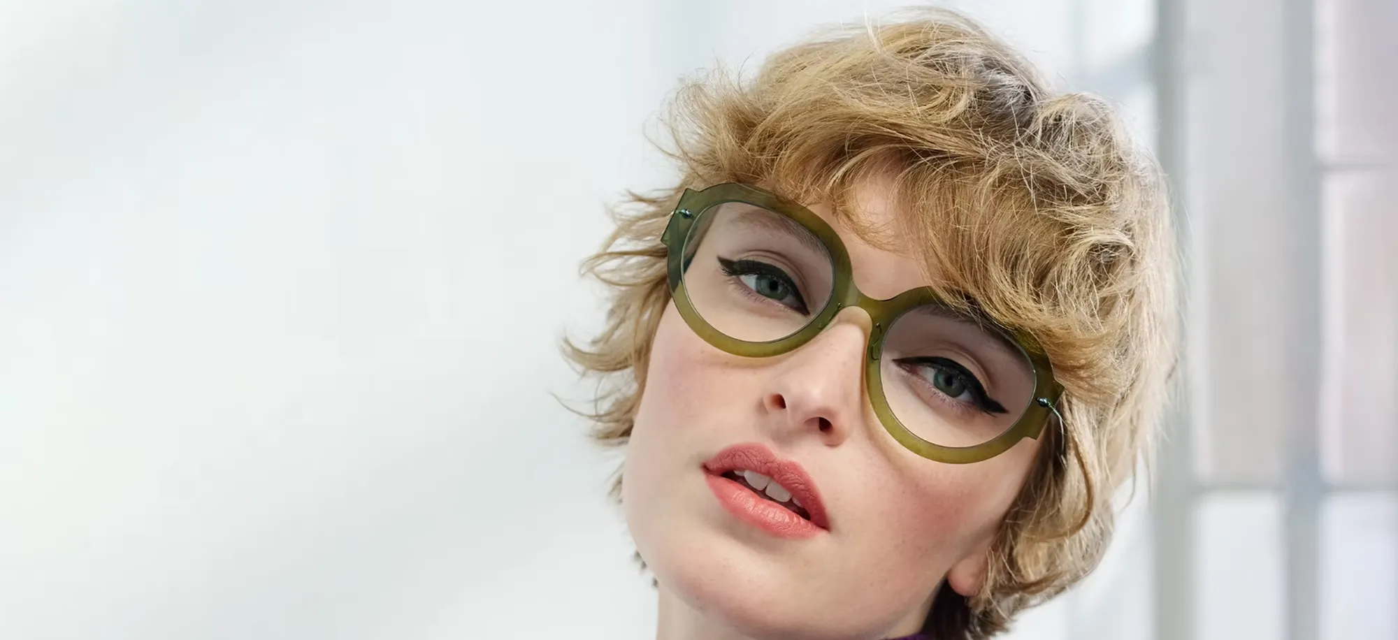 Close-up of a woman with short curly blonde hair wearing large round green glasses and subtle makeup.