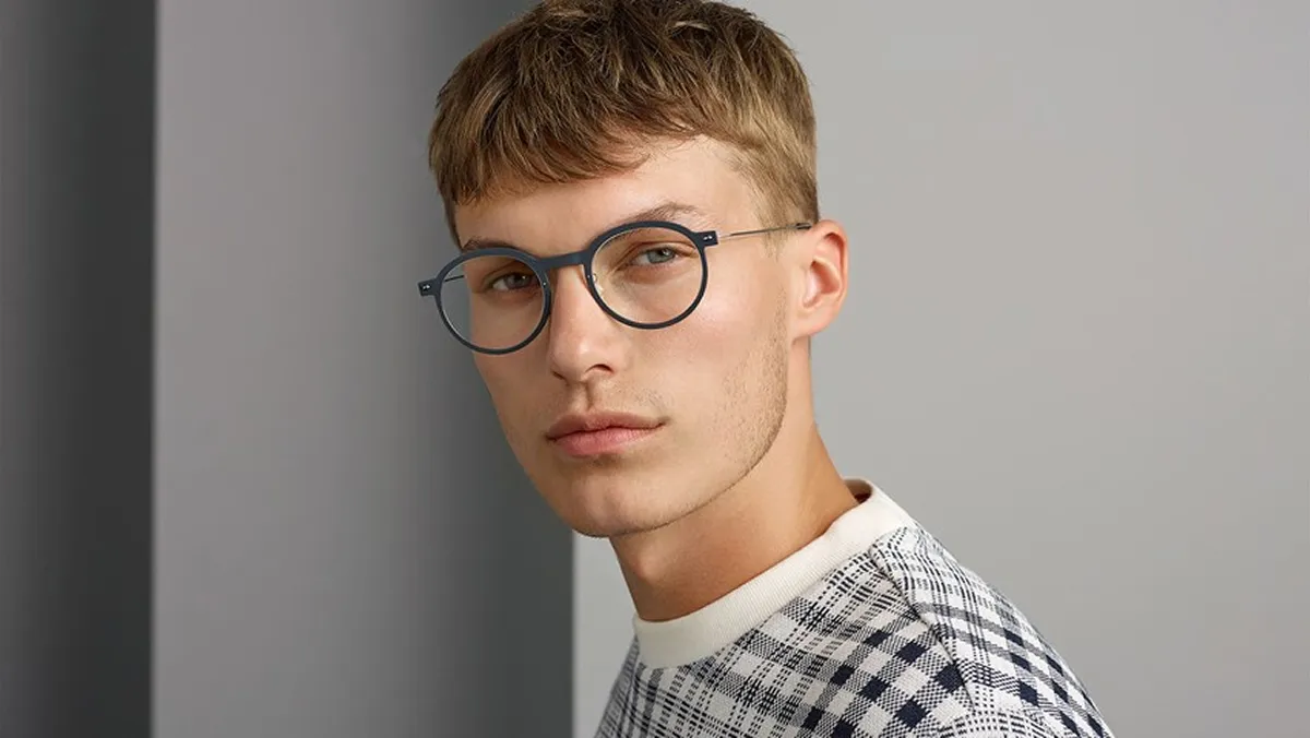 Young man wearing round gray glasses and a white and black checkered sweater, looking at the camera.