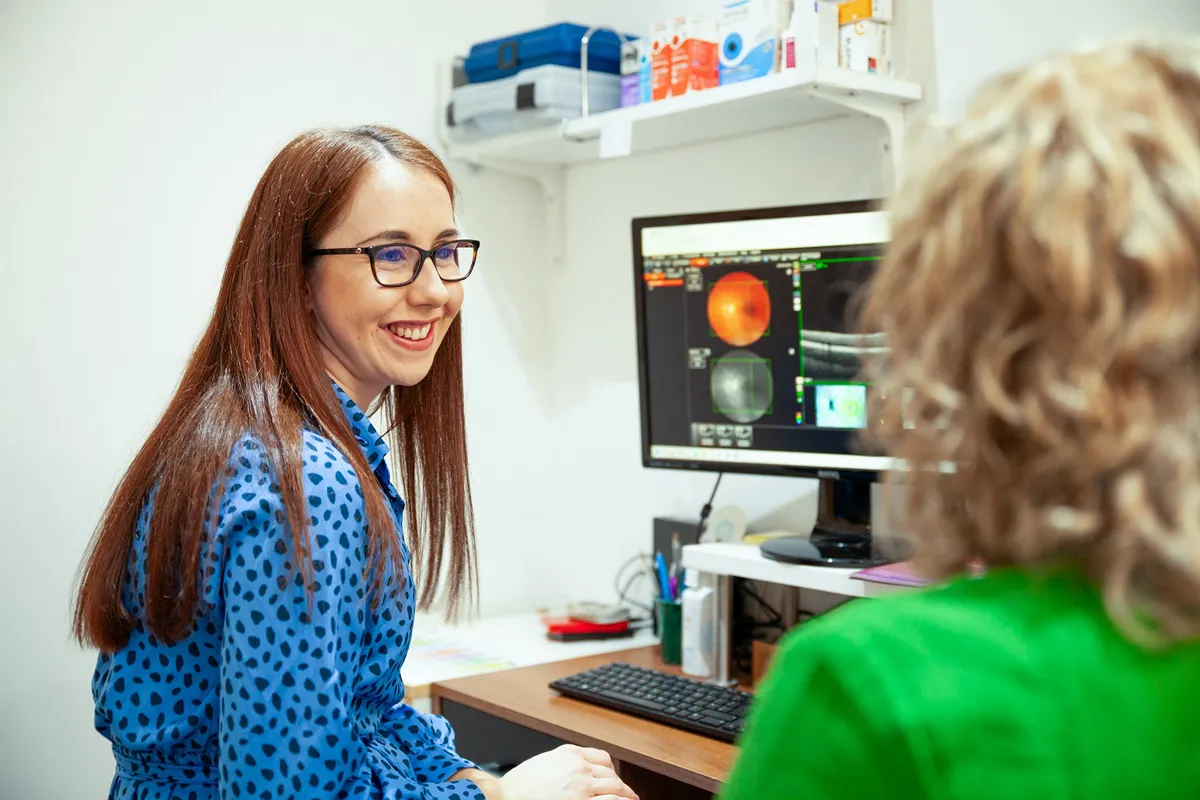 Female eye doctor wearing glasses smiling while consulting a patient in front of a computer showing eye scan images.