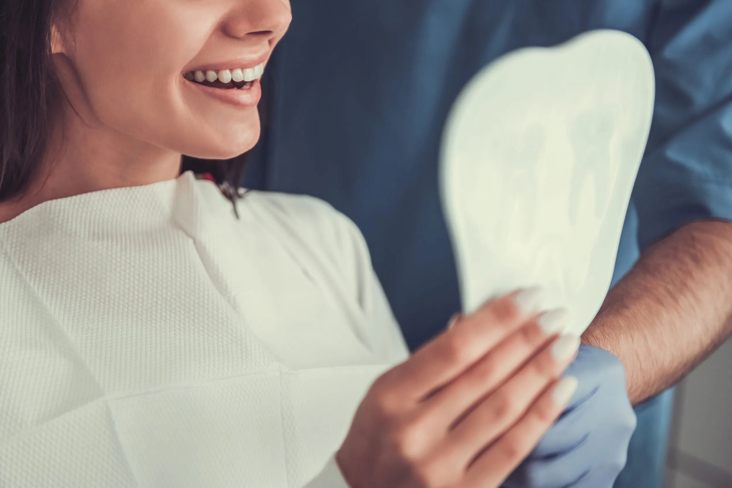 Smiling woman in dental bib holding a tooth-shaped mirror with a dental professional in blue gloves in the background.
