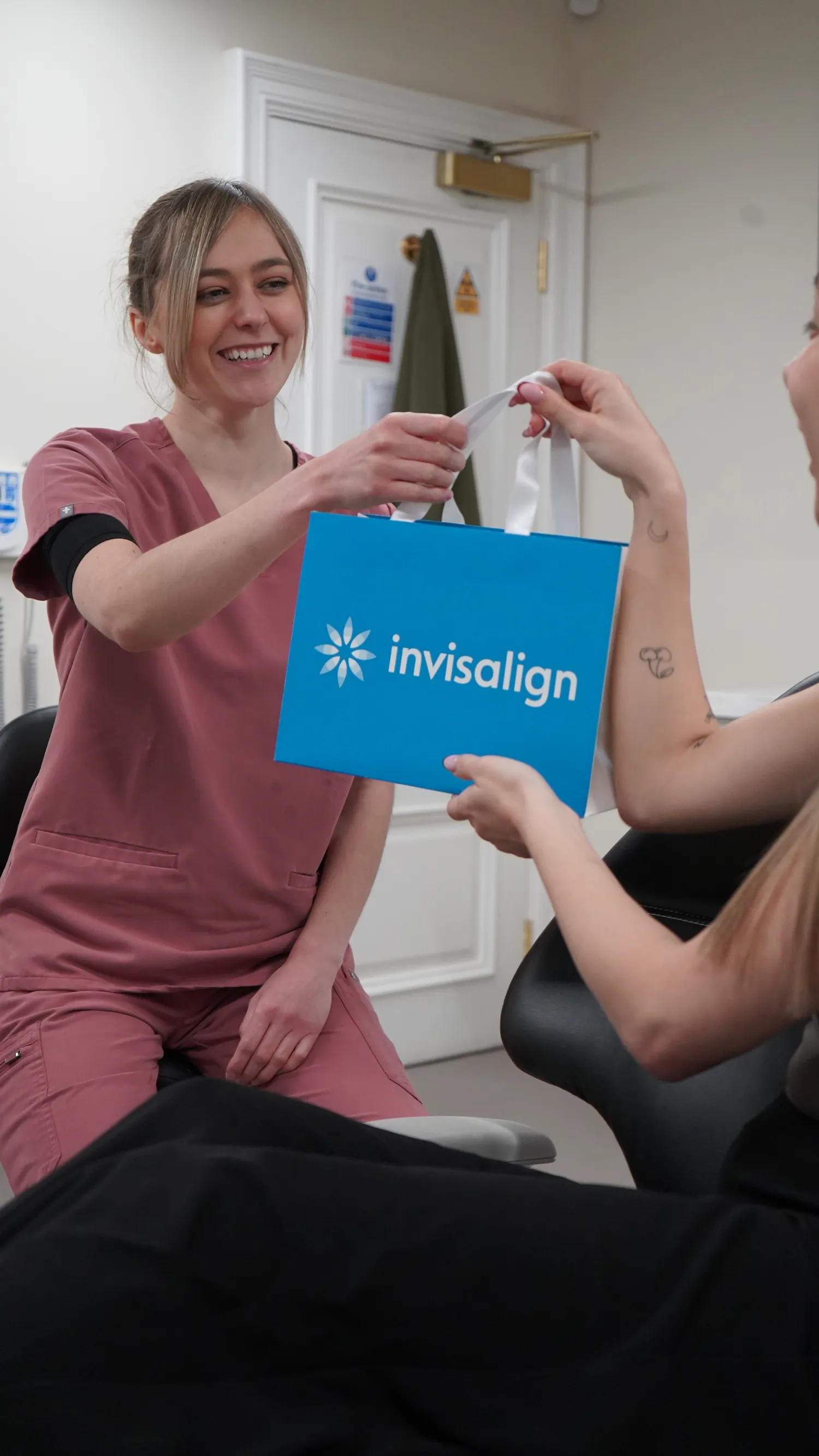 Smiling dental professional in pink scrubs hands a blue Invisalign bag to a seated patient.