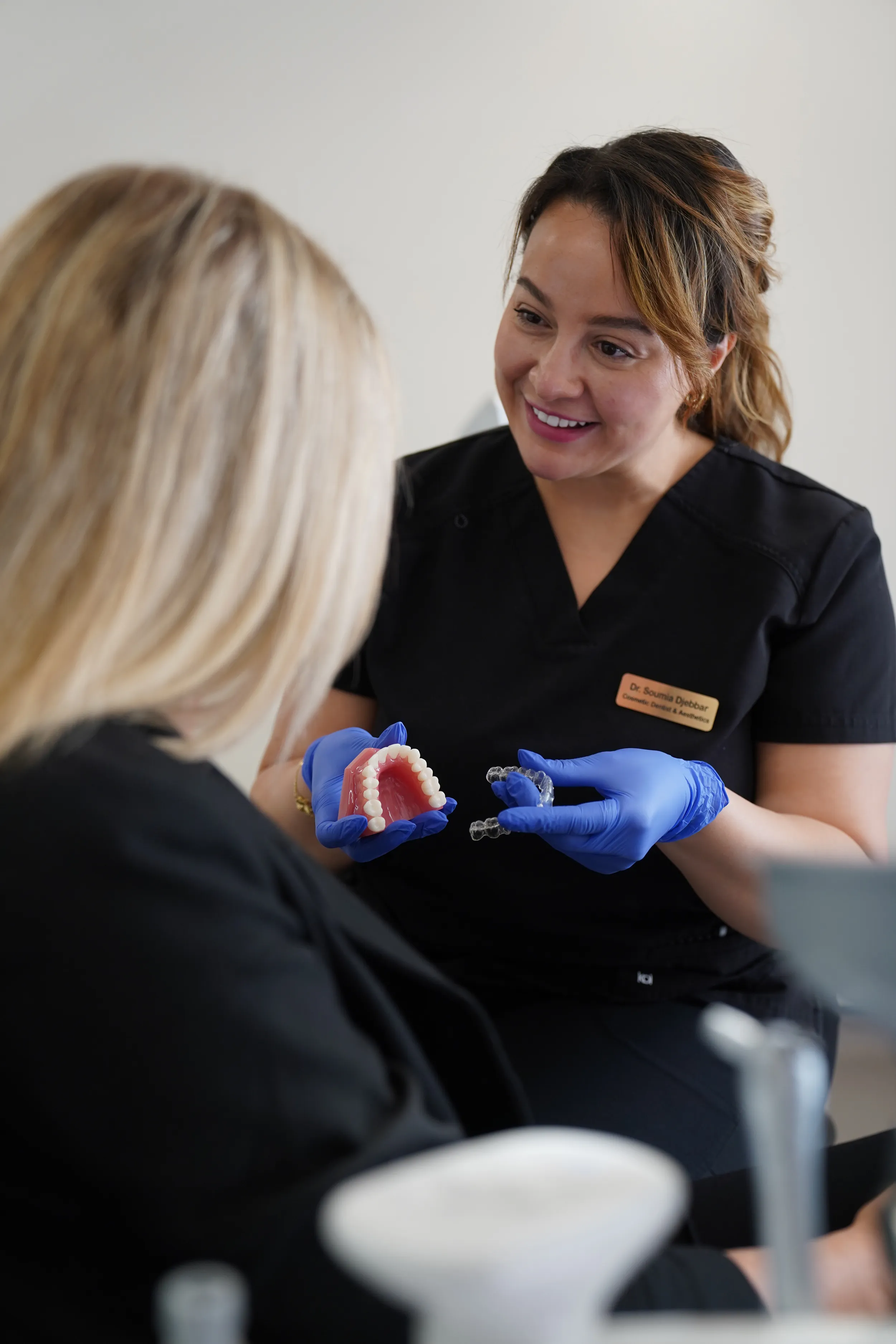Dentist wearing blue gloves shows a dental model and clear aligner to a seated patient.