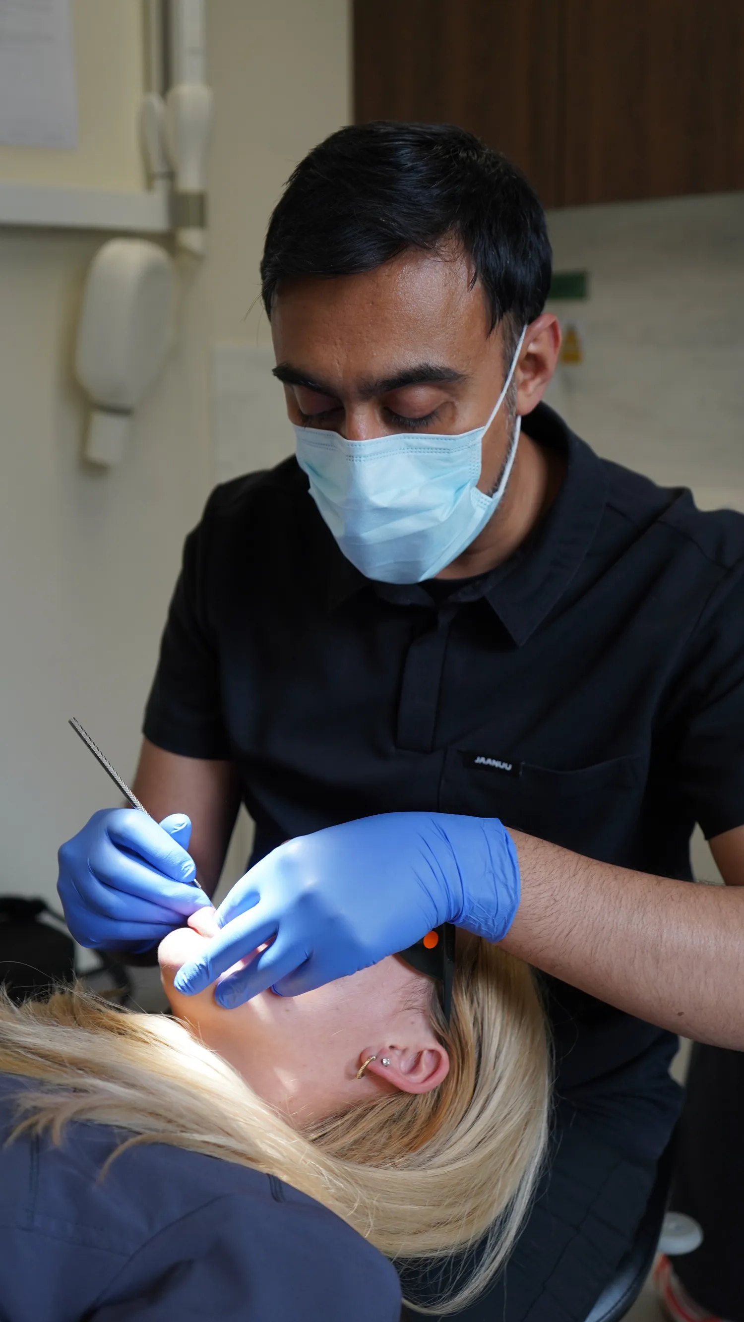 Dentist wearing a mask and blue gloves examining a patient's mouth with dental tools in a clinic.