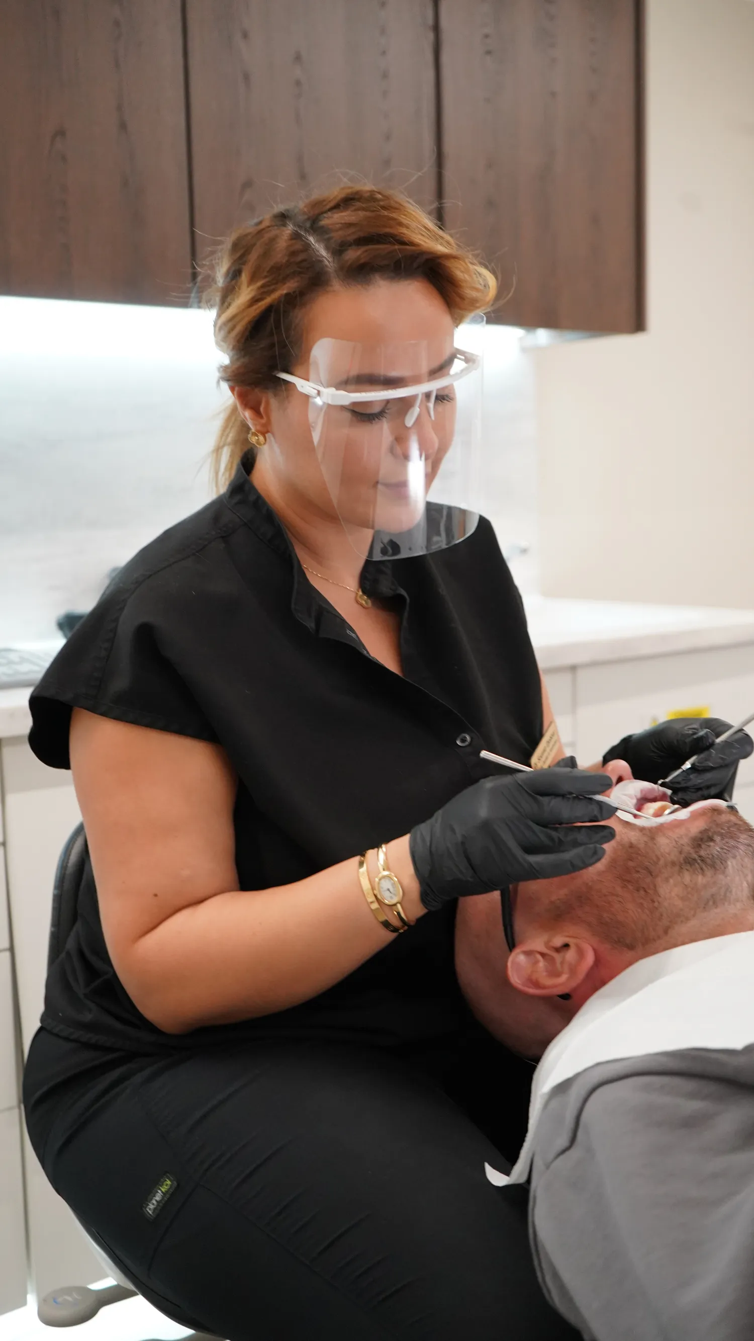 Dentist wearing a face shield and black gloves treating a male patient in a dental clinic.