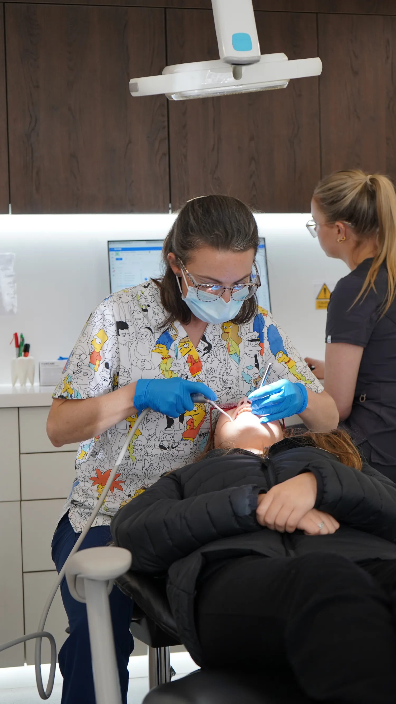 Dentist wearing glasses, a cartoon-themed scrub top, mask, and gloves performing a dental procedure on a patient lying in a dental chair.
