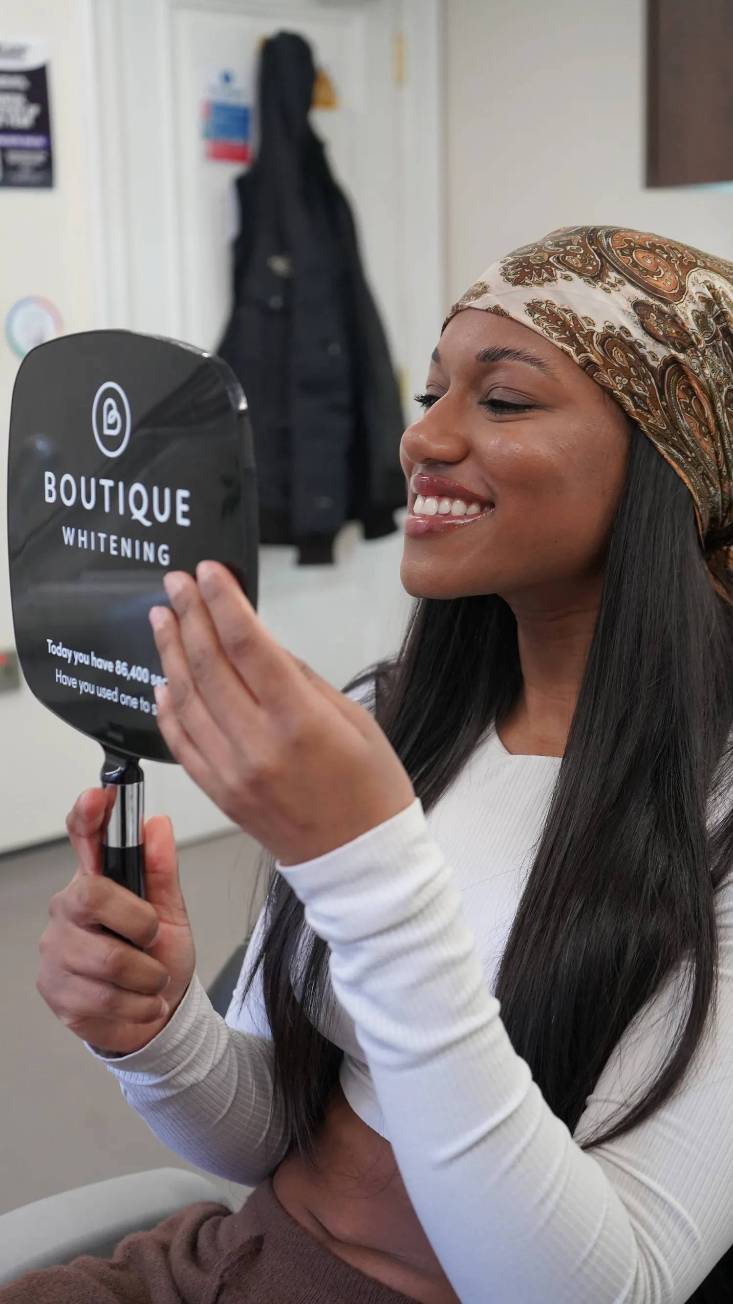 Woman wearing a patterned headscarf smiling and looking at her teeth in a Boutique Whitening handheld mirror.