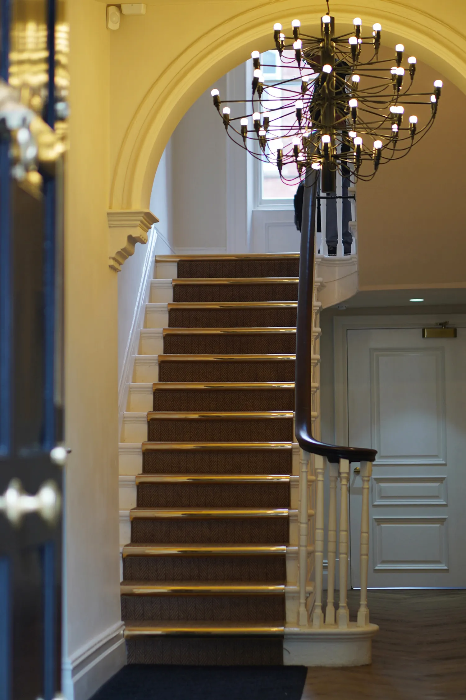 Indoor staircase with brown carpet and brass stair rods under a large archway with a modern chandelier hanging above.