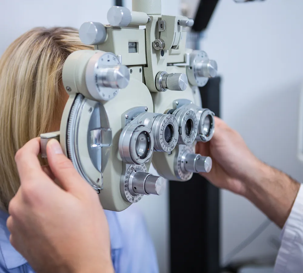 Close-up of an eye exam with a patient looking through a phoropter held by an optometrist adjusting the device's dials.